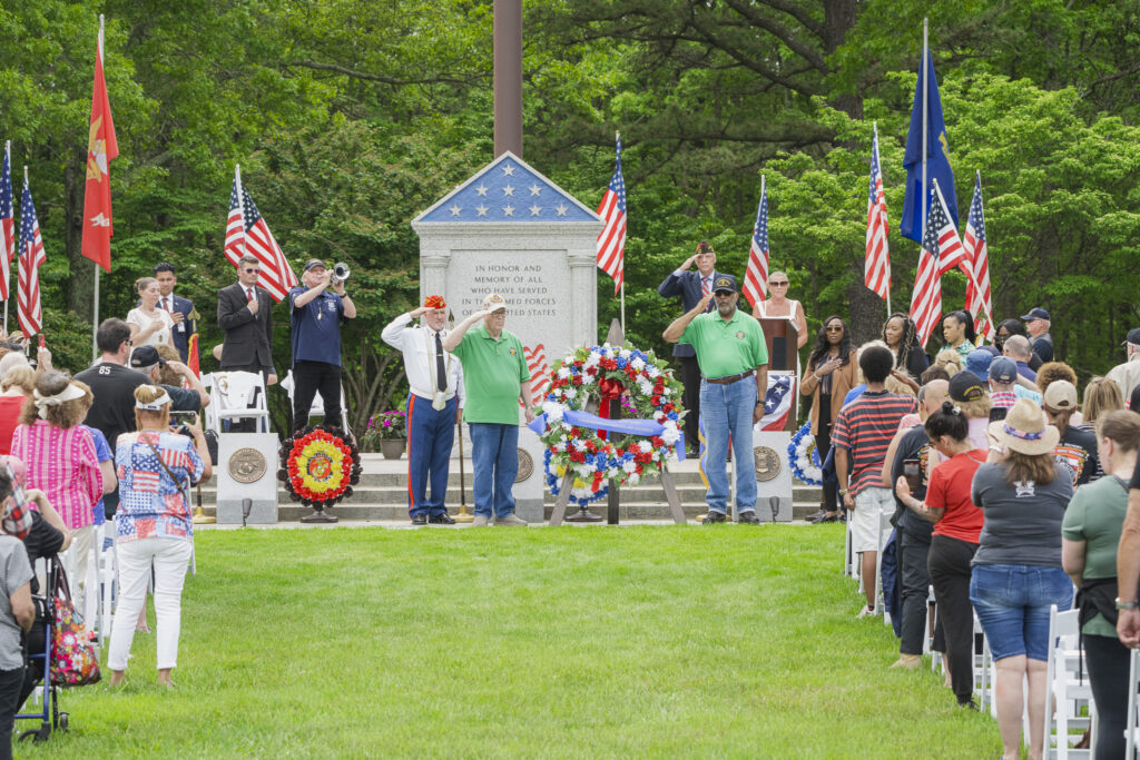 A Memorial Day commemoration ceremony where men stand at attention, saluting and a crowd sits in chairs. American flags wave throughout the property.