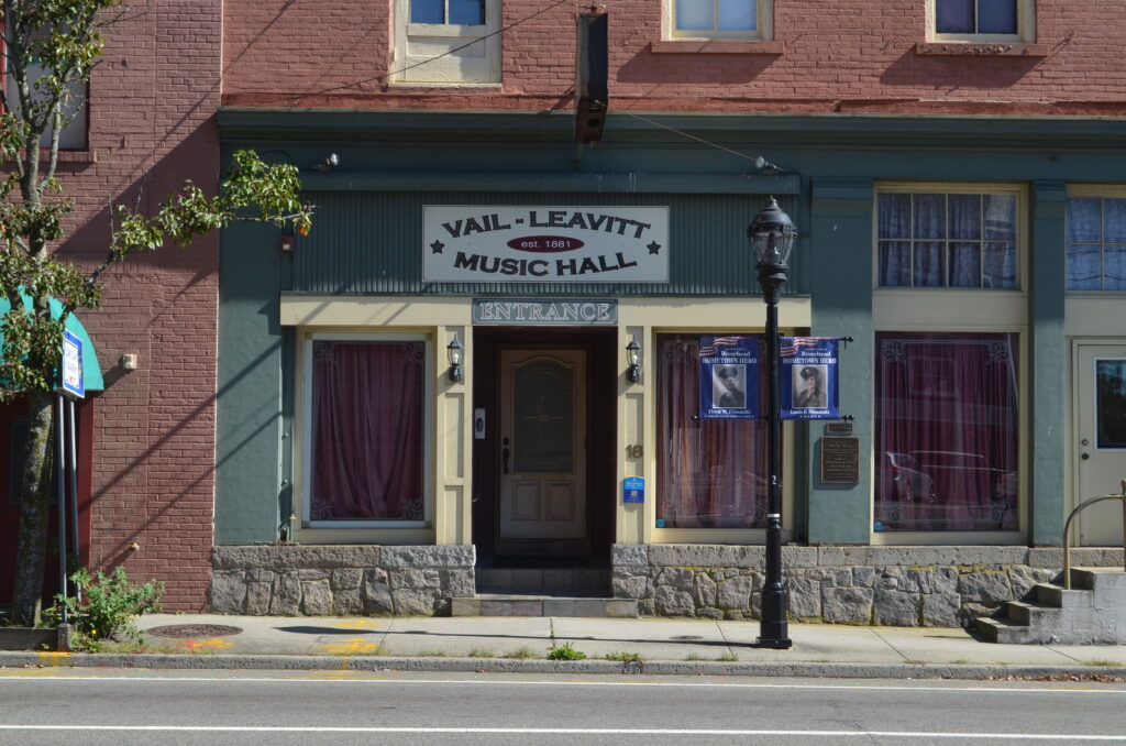 A picture of the outside of the Vail-Leavitt Music Hall in Riverhead. The building is brick, and the storefront itself to the entrance of the Vail-Leavitt is painted a muted green with cream trim. All of the windows are obscured with curtains.