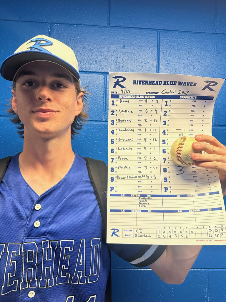 A high school baseball pitcher stands in front of a blue wall, holding up the baseball and blank score sheet from the perfect game he pitched.