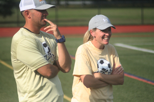 A man on the left and a woman on the right coach soccer from the sidelines.
