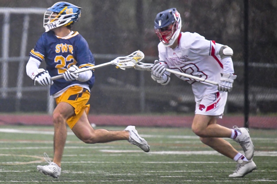 A Wildcats boys lacrosse player — in navy with yellow and white detailing — and an opposing player — dressed in a white uniform with red detailing — vie for the ball.