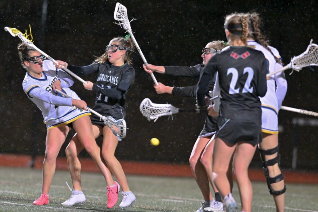 Two Shoreham-Wading River Wildcats girls lacrosse players — dressed in white uniforms with green and yellow trim — and two Westhampton girls lacrosse players — dressed in black uniforms with white and red detailing — vie for the ball at a rainy evening game.