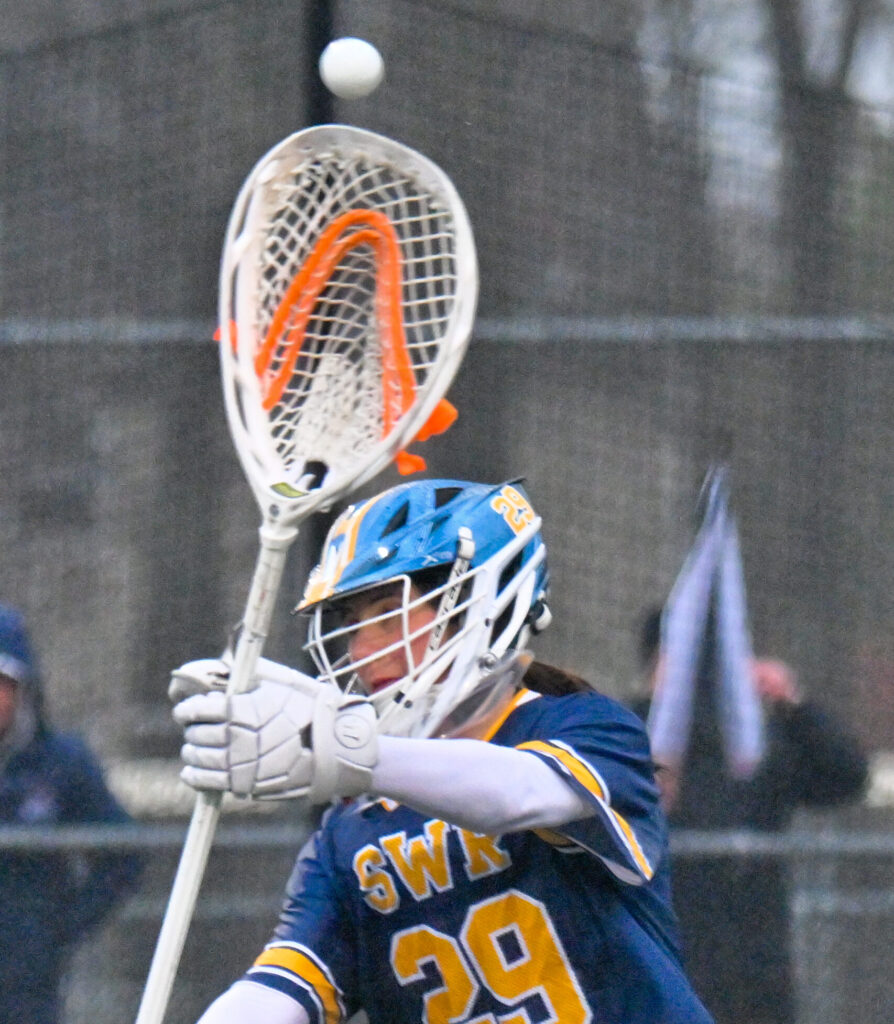 Senior Angelina Price, goaltender for the SWR lacrosse squad, throws the ball to one of her teammates. She wears the SWR blue uniform, complete with yellow and white detailing and trim. Her lacrosse stick's net is broad, fitting of the goaltender's net, and is white and orange.