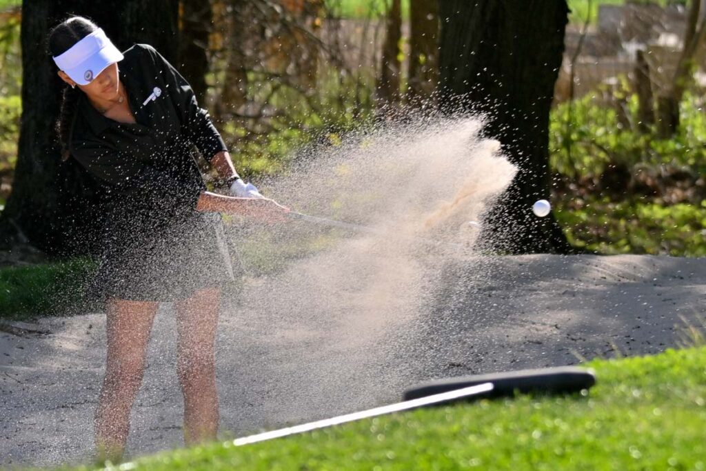 A girl in a white cap and blue golf shirt and skirt hits a golf ball out of a sand pit, blowing sand everywhere