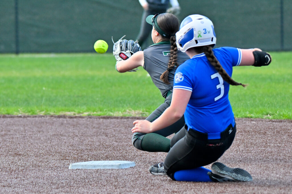 A softball player in a blue jersey, black pants and blue socks slides into a base as the ball comes to the opponent's mitt.