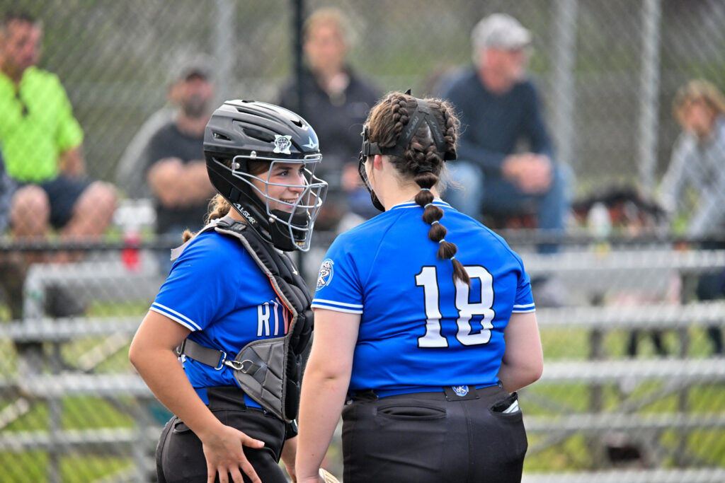 A softball pitcher and catcher in blue jerseys and black pants confer on the pitching mound.