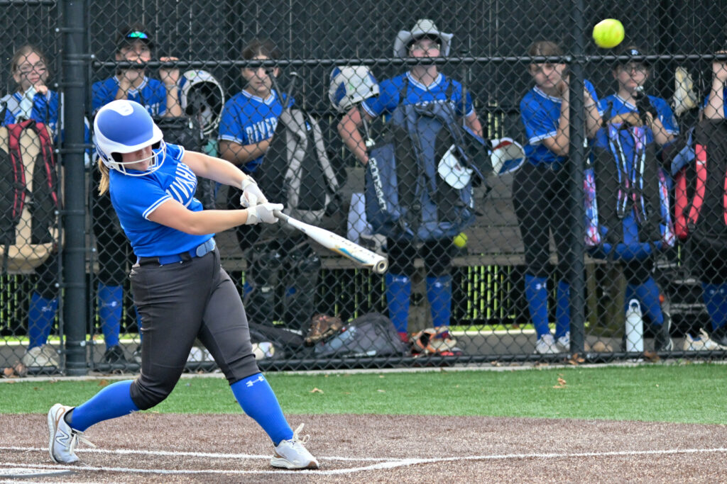 A softball pitcher in a blue jersey, black pants and blue socks hits the ball.